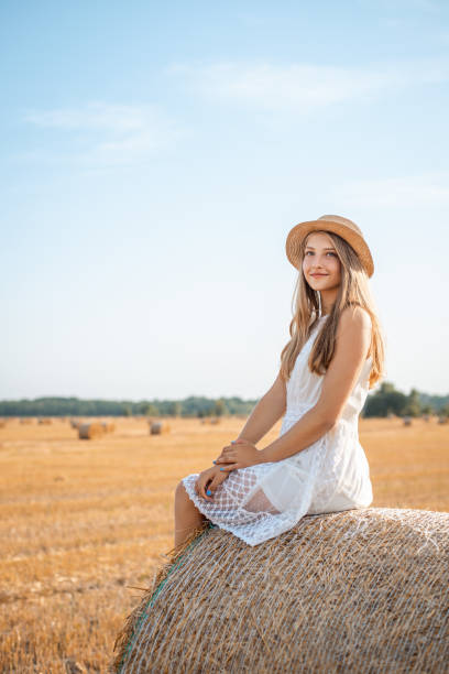 positive girl in a white dress and a straw hat sitting on a straw bale on the agricultural field - wheat sunset bale autumn imagens e fotografias de stock