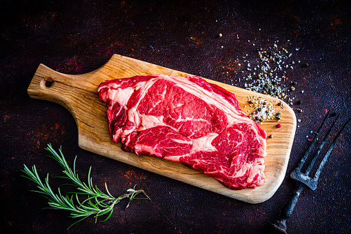 Fresh juicy raw Angus beef steak on a cutting board shot from above on brown background. Herbs and spices like rosemary, marine salt, peppercorns and chili peppers complete the composition. High resolution 42Mp studio digital capture taken with SONY A7rII and Zeiss Batis 40mm F2.0 CF lens
