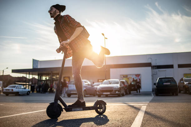 A young man on his e-scooter. Technology, ecological concept of mobility. A hipster young man riding an electric scooter. A modern young man with his electric scooter walks through the parking lot next to the shopping center while the sunset is behind him. push scooter stock pictures, royalty-free photos & images