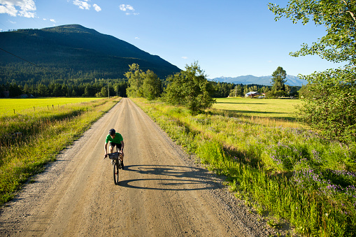 A male cyclist goes for a gravel road bicycle ride in British Columbia, Canada. His bicycle has bags strapped to the frame that carry extra clothes, food, and bike tools. His bicycle has a front suspension fork, disc brakes, and wheels and tires suited to riding on rough terrain.