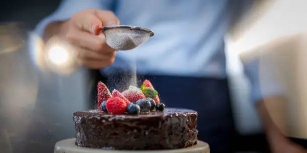 Man finishing tasty multi-tier cake. Male hand holding strainer and sprinkling top of cake with icing sugar. Homemade baked desserts for celebration or birthday concept.