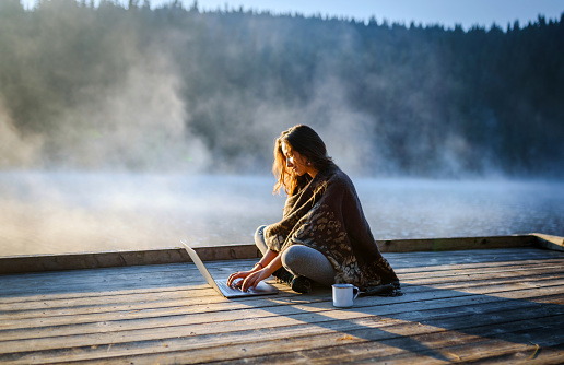 Woman Relaxing In the Nature And Using Technology. Side view of a beautiful woman relaxing near a lake with coffee and using laptop.