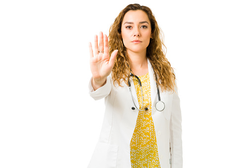 Hispanic female doctor making a hand gesture and telling someone to stop. Attractive medical specialist with a white coat preventing the spread of the virus