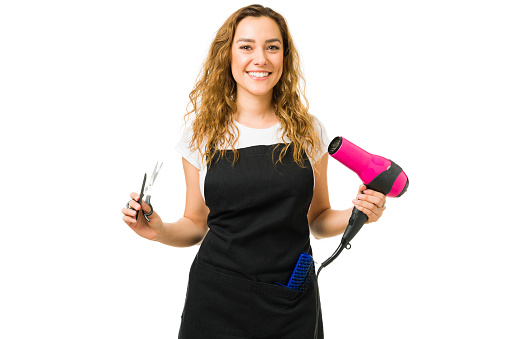 Happy attractive young woman smiling while making eye contact and holding a blow dryer with some scissors. Hair stylist working at a beauty salon