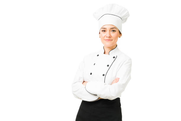 Beautiful female cooker smiling and posing in a white background Attractive young woman wearing a chef's uniform and hat. Female chef making eye contact with her arms crossed against a white background chefs whites stock pictures, royalty-free photos & images