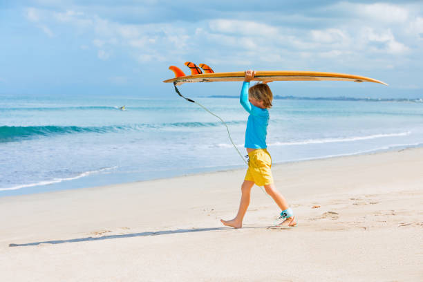 Young surfer learn to ride on surfboard on sea waves Happy surf boy - young surfer learn to ride on surfboard with fun on sea waves. Active family lifestyle, kids outdoor water sport lessons, swimming activity in surfing camp. Summer vacation with child raro stock pictures, royalty-free photos & images