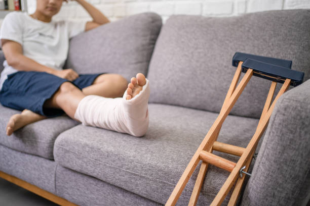 junger asiatischer mann mit gebrochenem bein auf dem sofa sitzend. nahaufnahme der krücke. - verletzung stock-fotos und bilder