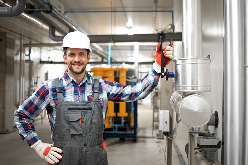 Portrait of factory engineer worker holding his arm on the valve and standing by heating or cooling pipeline system at boiler room. Gas installation maintenance.