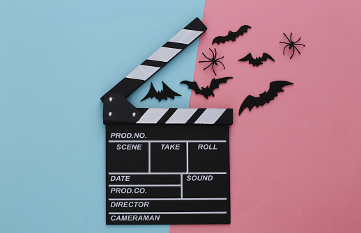 Horror movie, halloween theme. Movie clapperboard and flying decorative bats, spiders on pink blue pastel background. Top view, flat lay