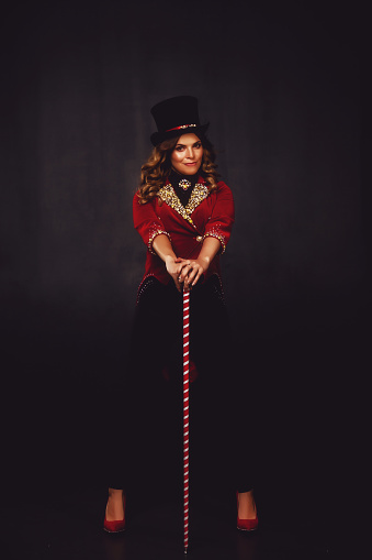Female magician illusionist circus with wand in theatrical clothes shows and smiles, on black background. Young woman actress in stage costume and top hat on head. Concept of performance. Copy space