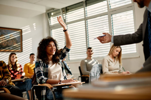 Happy black student raising her arm to answer teacher's question in the classroom. Happy African American female student raising arm to ask a question during lecture at university classroom. hand raised classroom student high school student stock pictures, royalty-free photos & images