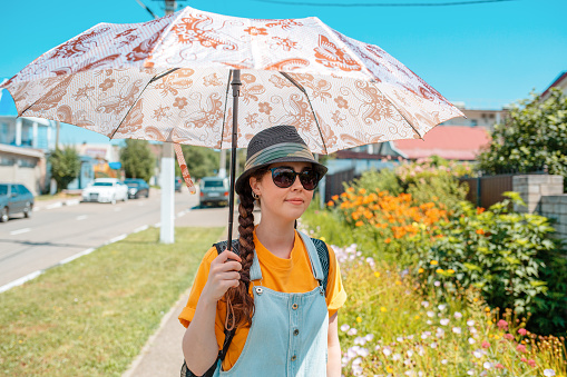 Portrait of woman stands on a green street in a straw hat, glasses and an umbrella, protecting herself from the sun. Outdoor. The concept of unwanted tanning.