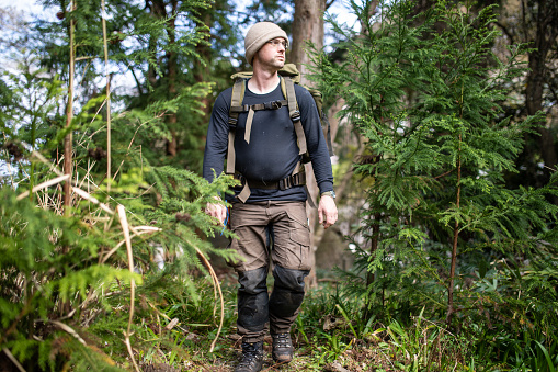 A Bushcraft survival man in outdoors gear walking through a woodland area.