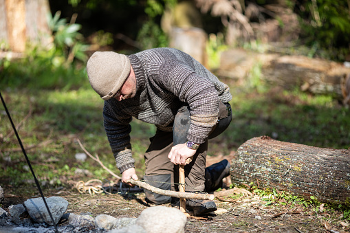 A bushcraft man starting a fire with a bow drill made in the woods. Smoke can be seen coming from the wood.