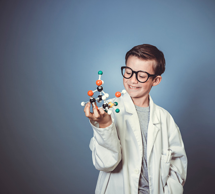 cool young boy is dressed as scientist with white lab coat and holding model of molecules in hand and wearing black glasses