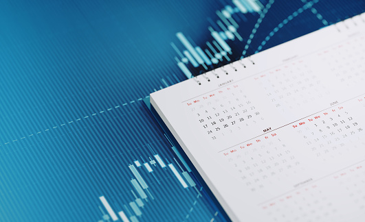 White calendar sitting on blue financial graph. Horizontal composition with selective focus and copy space. Investment, stock market data and financial planning concept.