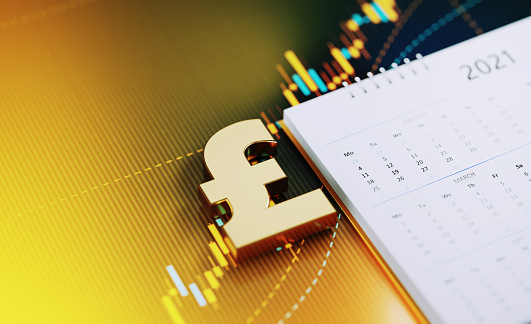 Gold colored British pound symbol sitting over a white 2021 calendar on yellow financial graph. Horizontal composition with selective focus and copy space. Investment, stock market data and financial planning concept.