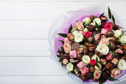 Beautiful bouquet of flowers and chocolate candies on white wooden background, top view. Space for text