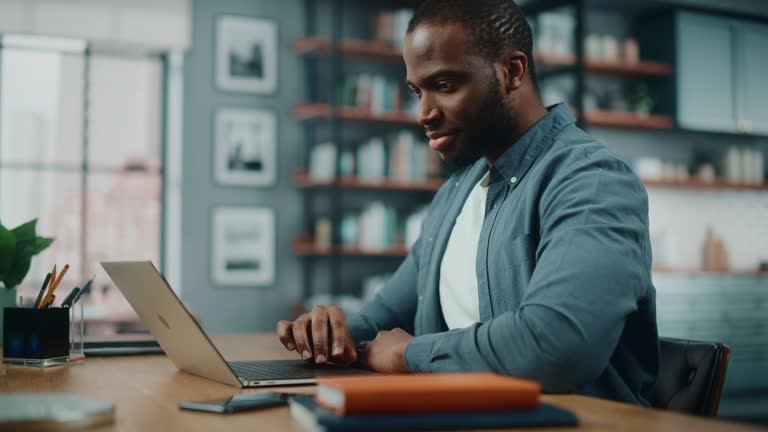 Handsome Black African American Man Working on Laptop Computer while Sitting Behind Desk in Cozy Living Room. Freelancer Working From Home. Browsing Internet, Using Social Network, Having Fun in Flat.