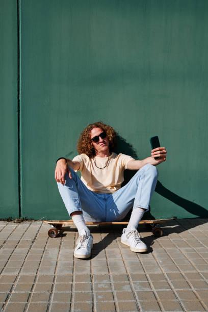 istock a young man using his smartphone and seated on his skateboard or longboard with a wall behind him 1312260476
