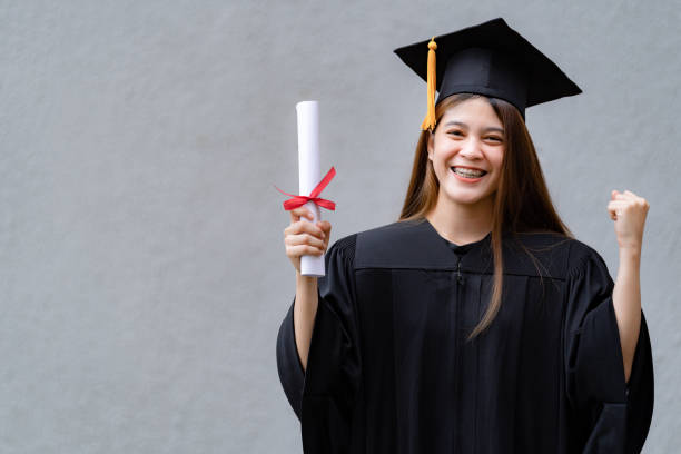 A young happy Asian woman university graduate in graduation gown and mortarboard holds a degree certificate celebrates education achievement in the university campus.  Education stock photo stock photo