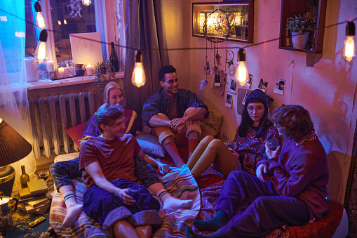 Group of young people sitting on the floor talking and having fun during domestic party in dark room