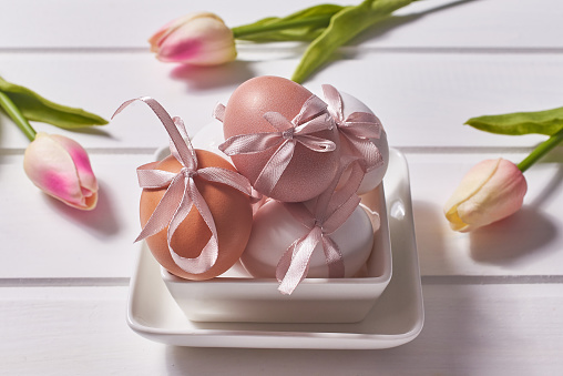 White and brown eggs with pink bows on a porcelain plate on a white wooden background and tulip flowers. Selective focus