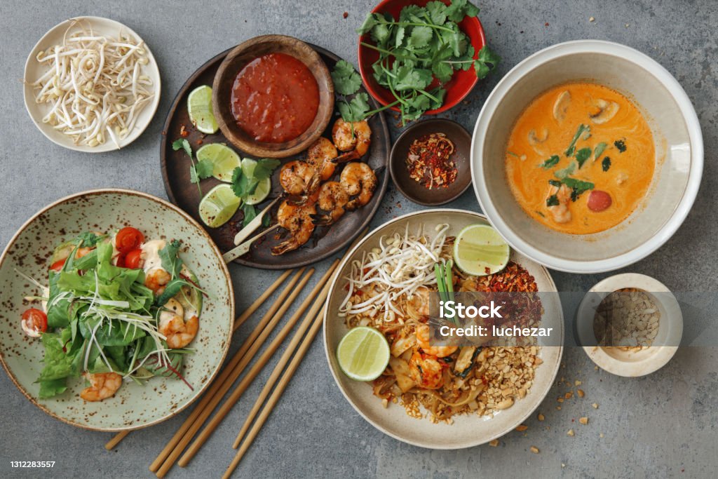 Classic Thai Food Dishes Authentic classic Pad Thai with shrimps. Thai shrimp satay with sweet chili sauce. Tom Yum soup with coconut milk (Tom Khaa). Spicy shrimp salad. Flat lay top-down composition on concrete background. Food Stock Photo