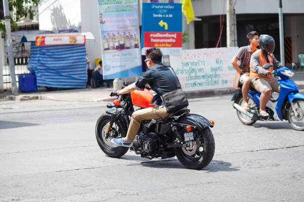 Thai man is driving Harley Davidson in Bangkok over a small junction in Ladprao. Model is 48 Fourty Eight. Man is not wearing a helmet. At right side a motorcycle  taxi is driving with customer