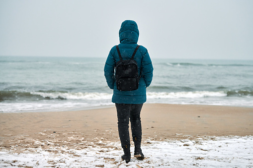 Back view of young woman tourist in green down jacket and with black backpack standing on sandy beach against background of sea horizon. Travel to sea under snowfall in winter.