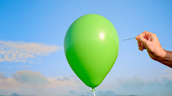 Close-up of male hand popping green balloon with pin against sky.