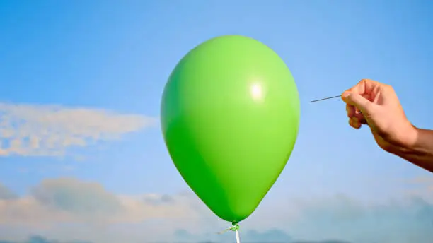 Close-up of male hand popping green balloon with pin against sky.