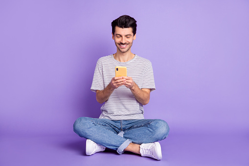 Photo of cheerful guy hold phone look screen beaming smile wear striped t-shirt jeans sneakers isolated violet background.