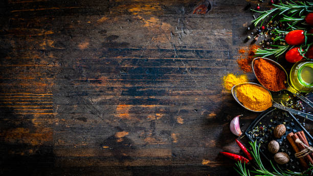 Cooking backgrounds: overhead view of multi colored spices and herbs arranged at the right of a dark wooden table making a border and leaving useful copy space for text and/or logo. The composition includes rosemary twigs, tomatoes, olive oil, paprika, turmeric nutmeg, cinnamon, garlic, chili pepper, marine salt and peppercorns. High resolution 35,5Mp studio digital capture taken with Sony A7rII and Sony FE 90mm f2.8 macro G OSS lens
