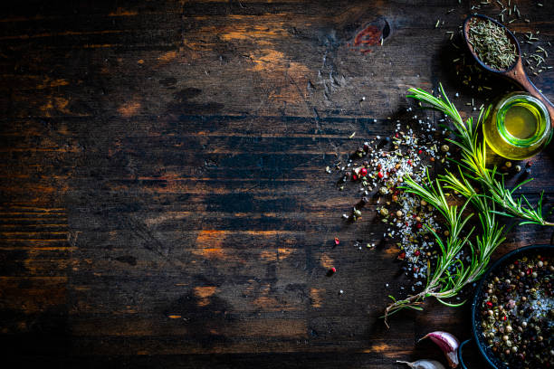 Culinary border of rosemary, salt, pepper and oil Culinary background: overhead view of rosemary twigs, marine salt, pepper, olive oil container arranged at the right of a dark wooden table making a border and leaving useful copy space for text and/or logo. Predominant colors are dark brown and green. High resolution 42Mp studio digital capture taken with Sony A7rII and Sony FE 90mm f2.8 macro G OSS lens cooking with garlic stock pictures, royalty-free photos & images