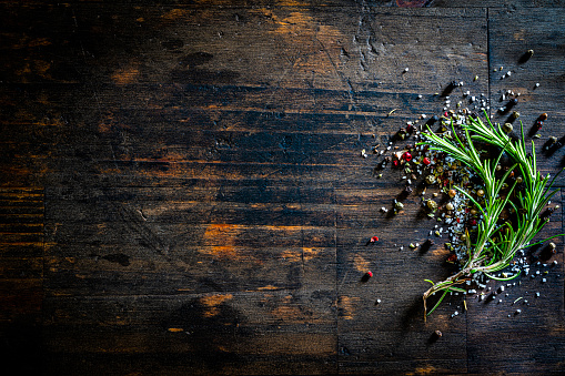 Cooking backgrounds: marine salt and mixed peppercorns scattered at the right of a dark brown wooden table. A fresh rosemary twig is on the salt and pepper. Copy space available for text and/or logo. Predominant colors are green and brown. High resolution 42Mp studio digital capture taken with Sony A7rII and Sony FE 90mm f2.8 macro G OSS lens