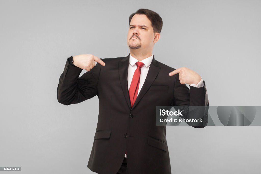 A very proud man shows his fingers and boasts A very proud man shows his fingers and boasts. indoor studio shot. isolated on gray background. handsome businessman with black suit, red tie and mustache looking away. Arrogance Stock Photo