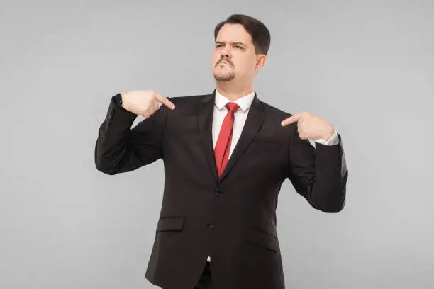 A very proud man shows his fingers and boasts. indoor studio shot. isolated on gray background. handsome businessman with black suit, red tie and mustache looking away.