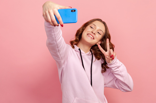 Portrait of happy teen girl in hoodie showing victory gesture with fingers on smartphone camera and sincerely smiling, vlog or selfie. Indoor studio shot isolated on pink background