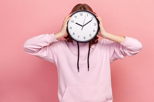 Portrait of young brunette female in hoodie holding wall clock hiding her face, time management, schedule and meeting appointment. Indoor studio shot isolated on pink background