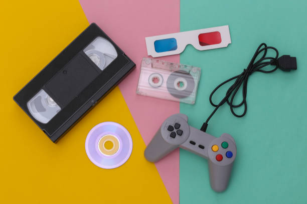 istock Wired retro gamepad, anaglyph 3D glasses, audio and video cassette, CD's on colored background. Retro Attributes. Top view 1312310656