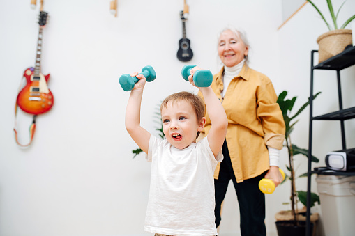 Tired granny doing phisical exercises with her grandson, trying to encourage him to stay fit. They are lifting dumbbells overhead. Boy is very enthusiastic and engaged.