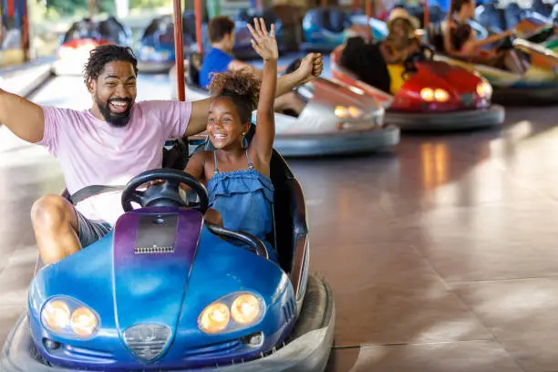 A Cheerful Family of African-American Ethnicity is Spending the Time Together and Riding in Bumper Car in Large Amusement Park. Excited Little Girl with Curly Hair is Enjoying the Time with her Father.
