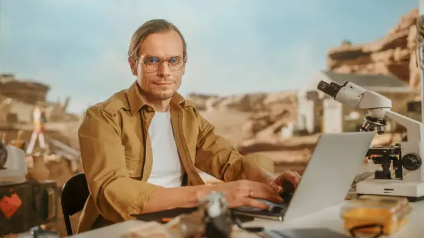 Photo of Archeological Digging Site: Portrait of Great Male Archaeologist Doing Research, Using Laptop, Looks at the Camera Smilingly, Analysing Fossil Remains or Ancient Civilization Culture Artifacts