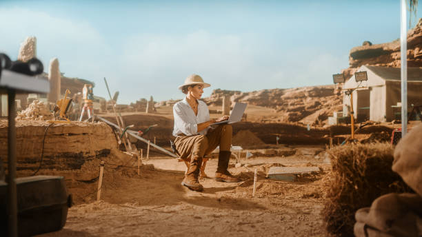istock Archeological Digging Site: Great Influencial Female Archaeologist Doing Research, Using Laptop, Analysing Unearthed Ancient Civilization Culture Artifacts Laying Nearby. Famous Historian Work 1312318135
