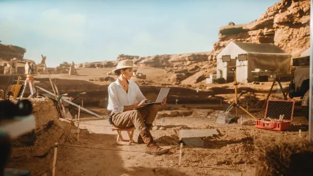 Archeological Digging Site: Great Influencial Female Archaeologist Doing Research, Using Laptop, Analysing Unearthed Ancient Civilization Culture Artifacts Laying Nearby. Famous Historian Work