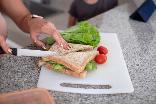 Close-up shot of an unrecognizable female hands making a vegetarian sandwich with lettuce and tomato for her son breakfast. She's cutting the sandwich in half on a cutting board with a kitchen knife while her son standing next to her.