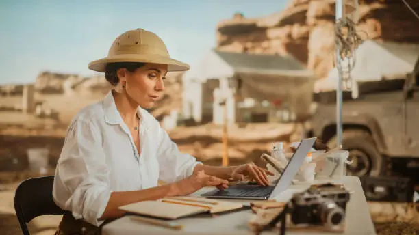 Photo of Archeological Digging Site: Great Female Archaeologist Doing Research, Using Laptop, Analysing Fossil Remains, Ancient Civilization Culture Artifacts. Team of Historians work on Excavation Site