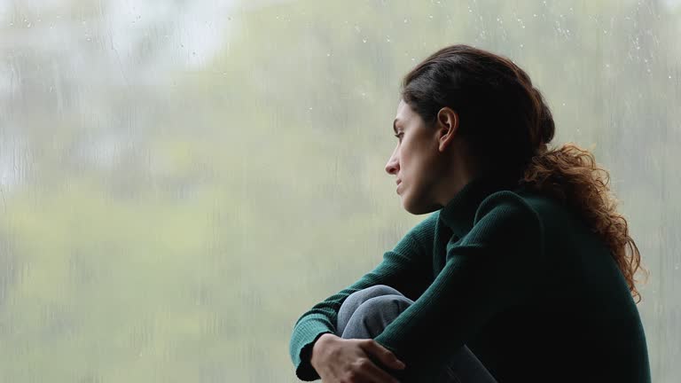 Sad melancholic young woman sitting on windowsill, watching rainy weather.