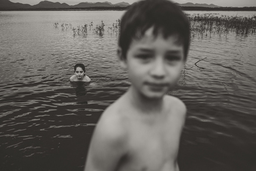 A black and white photography with two kids (a boy unfocused in the foreground and a girl in the background) into a calm lake, at Iraí Reservoir, in the city of Pinhais, State of Paraná, Brazil